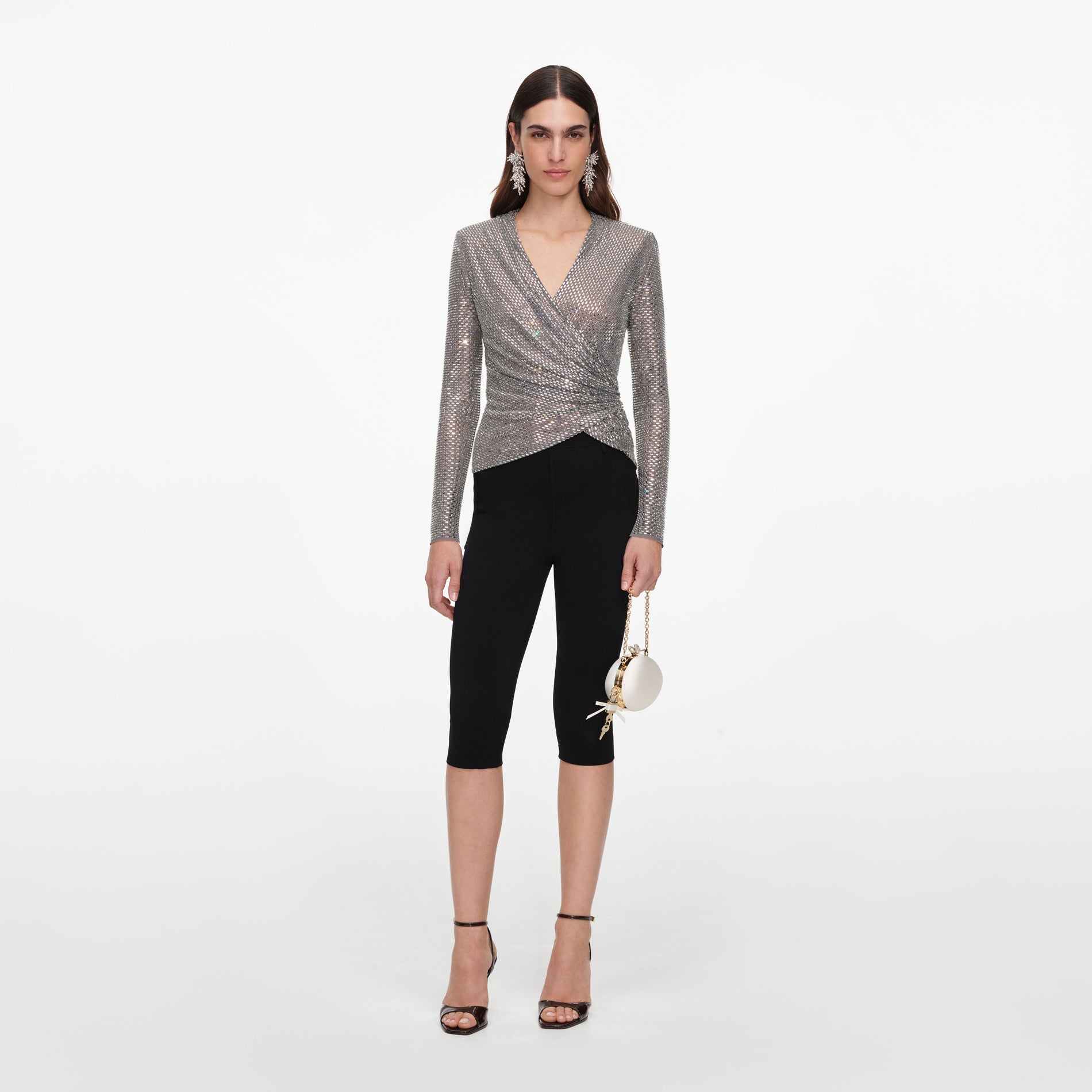 Woman wearing a silver sequin wrap top, black capri pants, black high-heeled sandals, and holding a white round purse