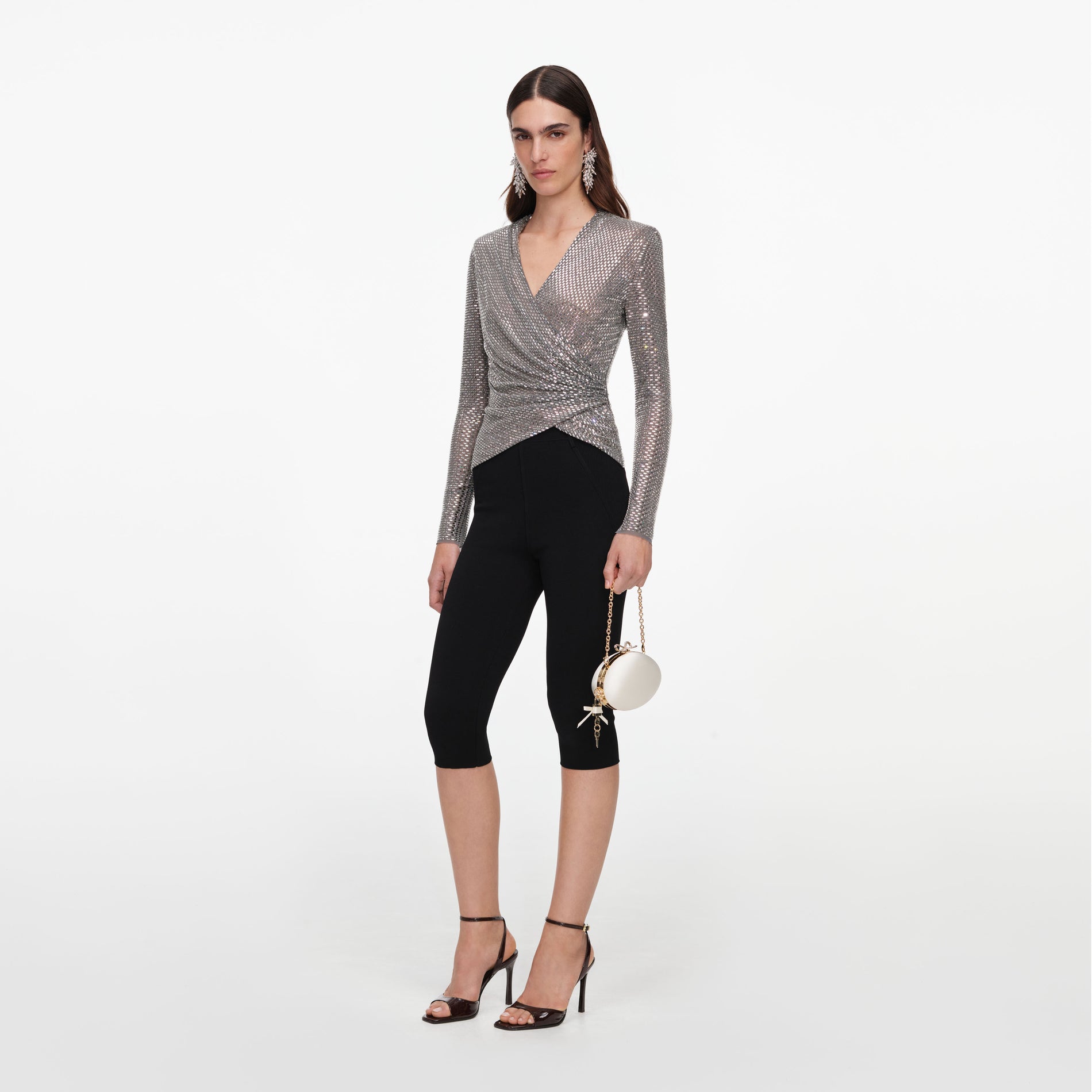 Woman in a silver sequin wrap top, black capri pants, dark brown high heels, and a white round clutch