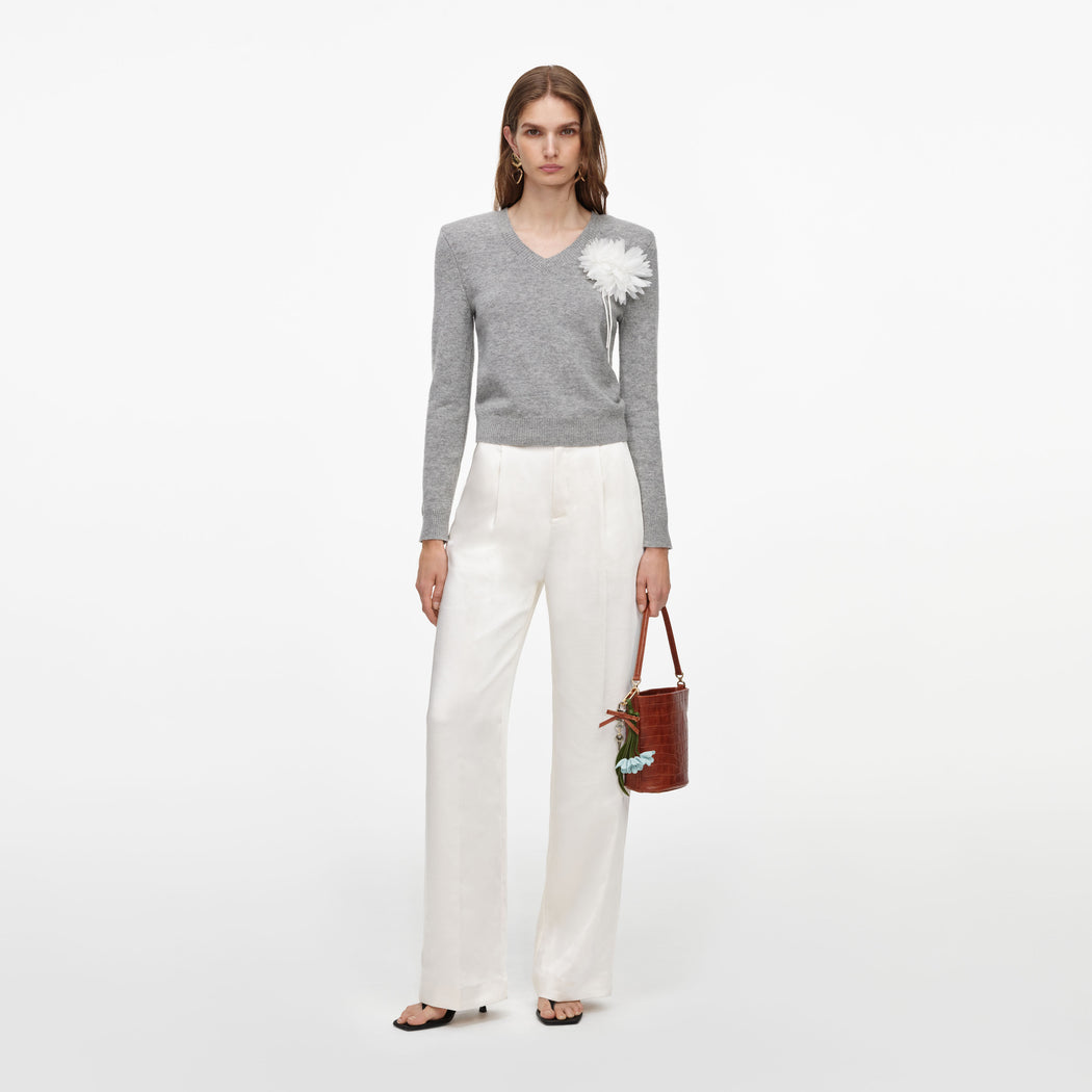 Woman wearing a grey v-neck sweater with a white flower brooch, white wide-leg trousers, and black sandals