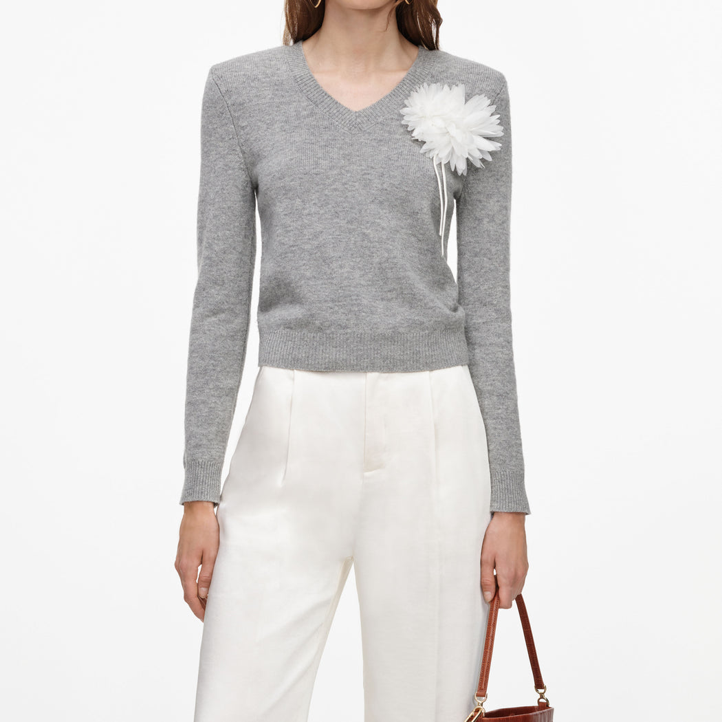 Woman wearing a grey v-neck sweater with a white floral embellishment and white trousers
