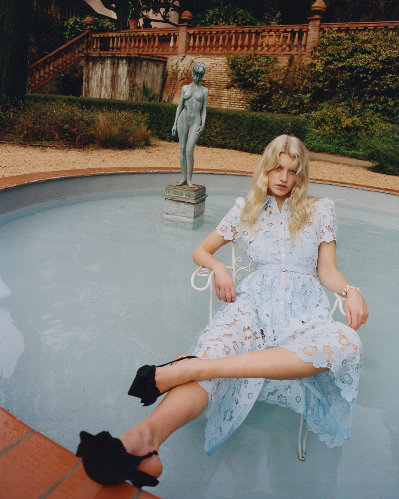 Woman in a blue lace dress sitting by a pool with a statue in the background.